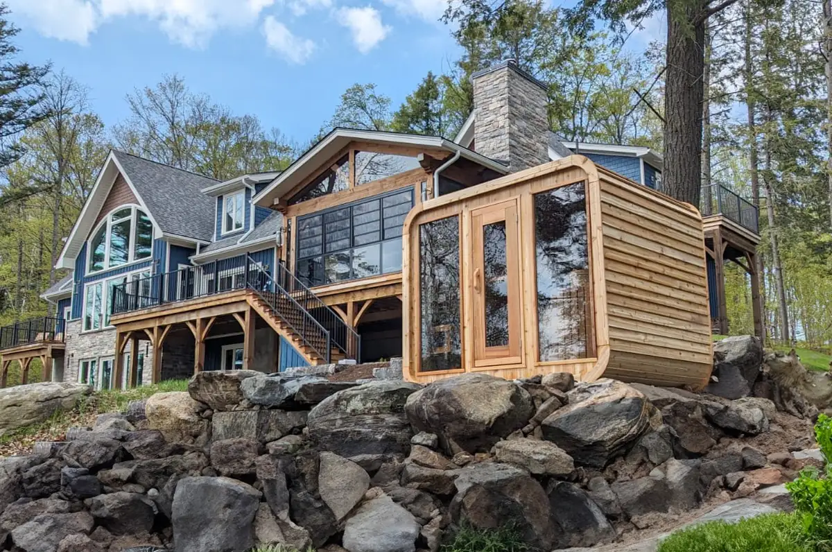 Dundalk LeisureCraft Luna Sauna in red cedar with a full glass front, set on a rock ledge beside a lakeside home